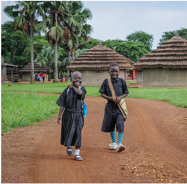 Two children walking down a dirt road with huts in the background near a Christian school.