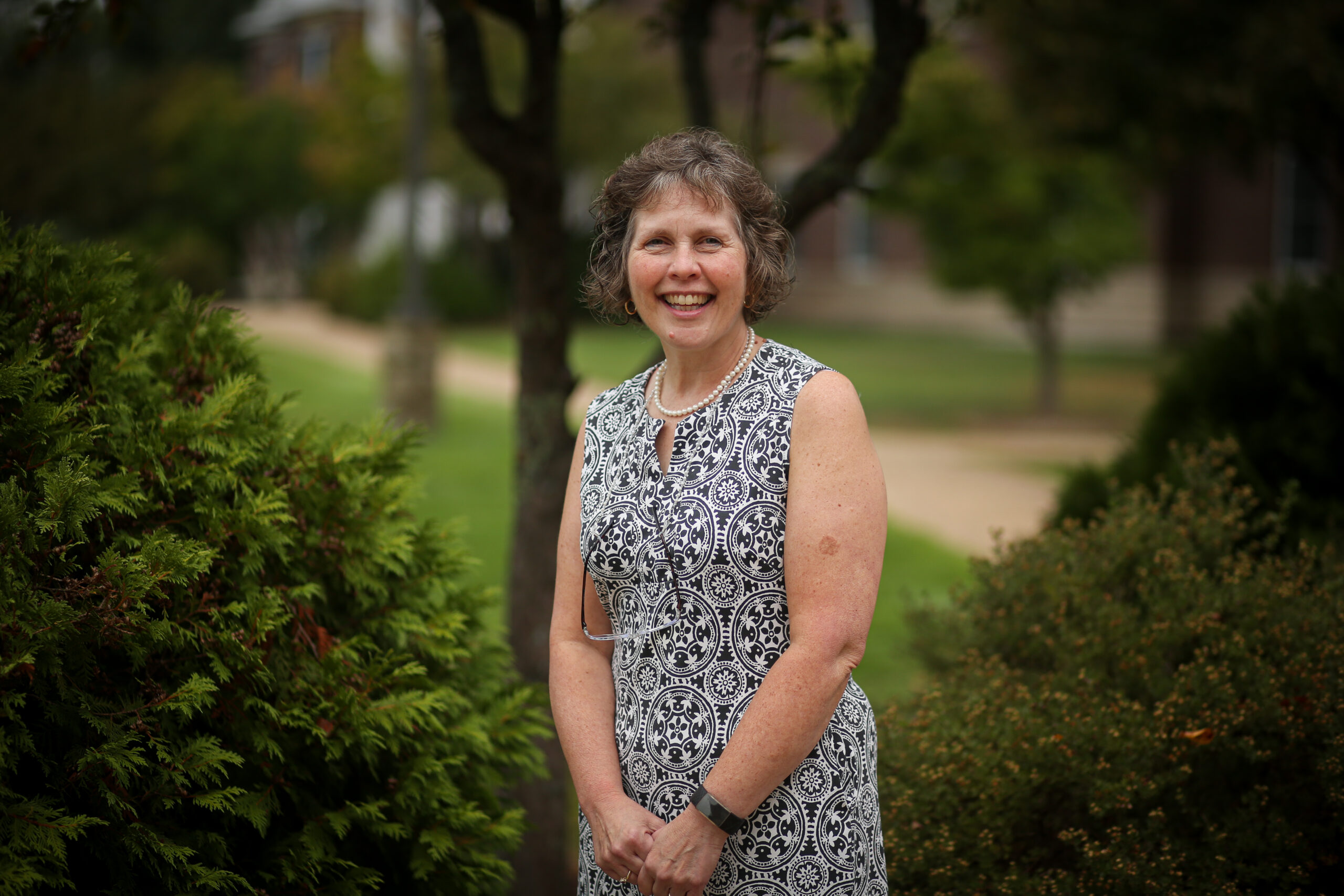 A smiling woman in a dress standing in front of trees, representing a Christian school.
