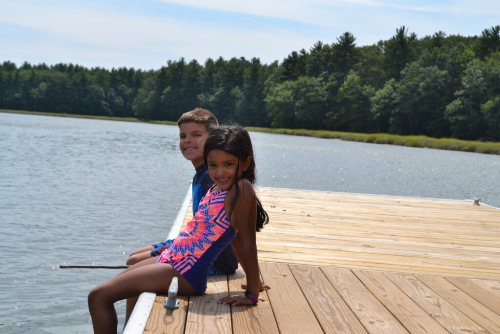Two children from a Christian school sitting on a dock near a body of water.