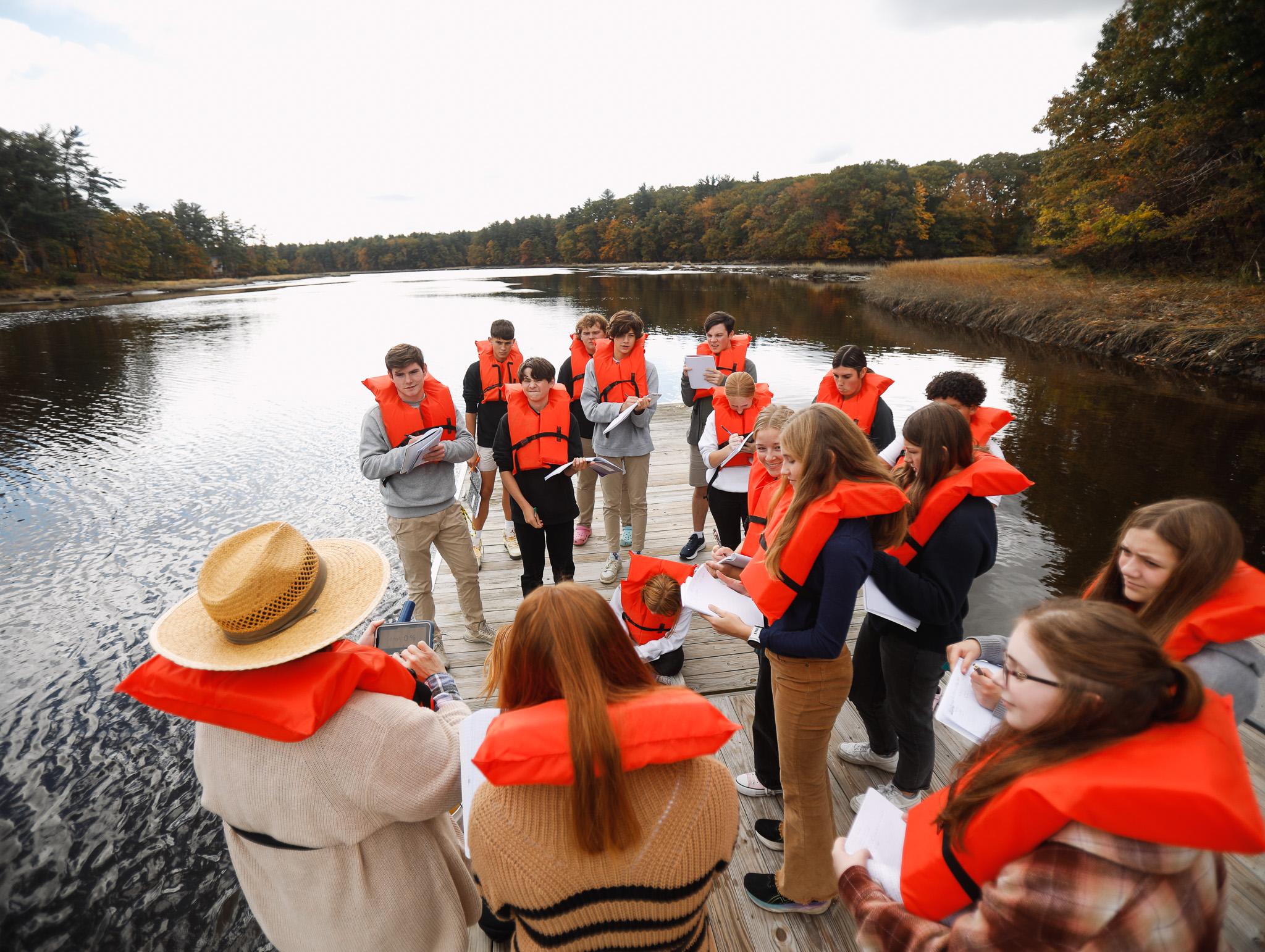 A group of people wearing orange life jackets stand on a dock by a body of water, engaged in a discussion or activity. Trees with autumn foliage are visible in the background.