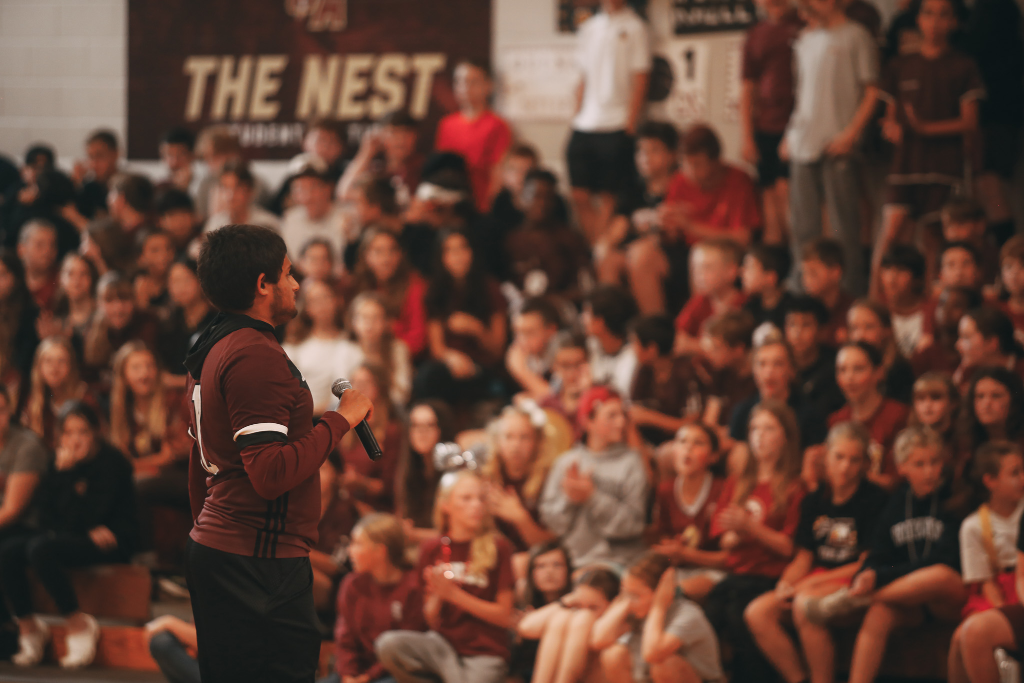 A person stands with a microphone facing a seated crowd in a gymnasium with "The Nest" sign in the background.