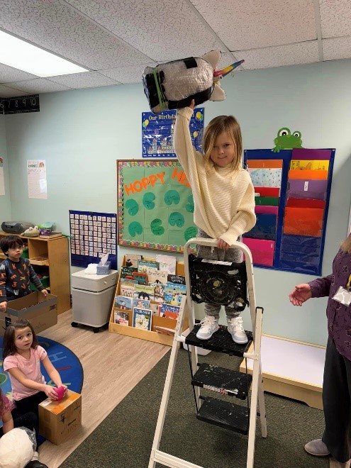 A little girl standing on a step stool in a classroom.