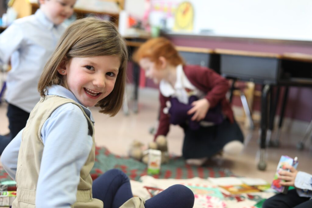 A young girl smiling at the camera while sitting on the floor, with other children playing in the background of a classroom.