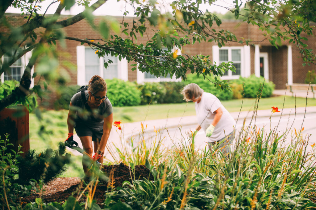 Two women gardening, one digging with a trowel near flowers, in front of a residential building.