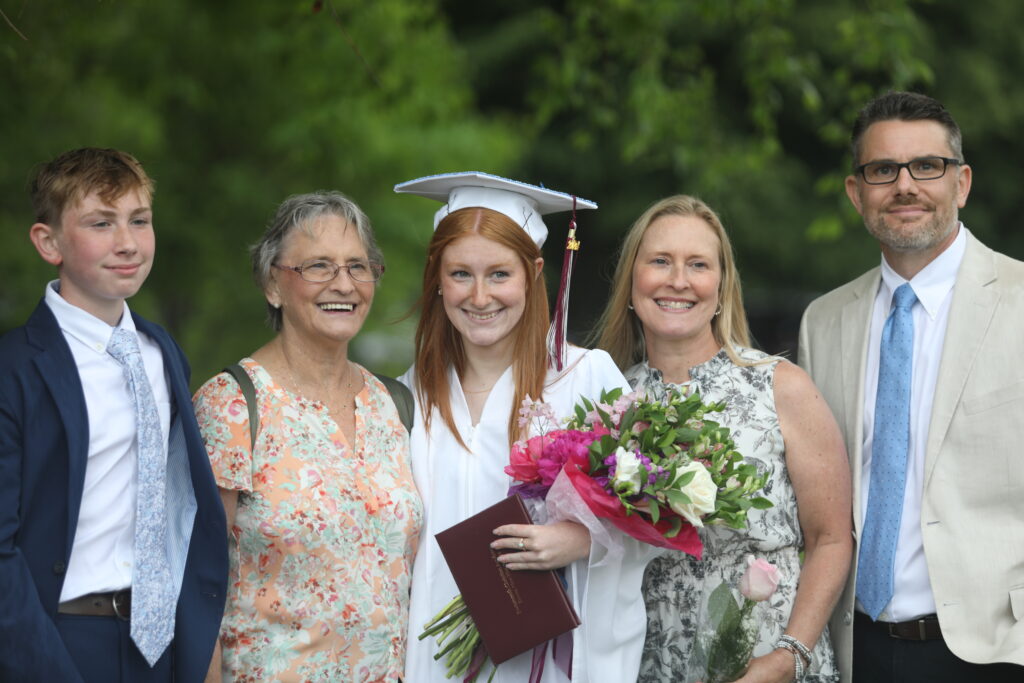 A graduate in a white cap and gown holds a diploma and flowers, standing with four people outdoors.