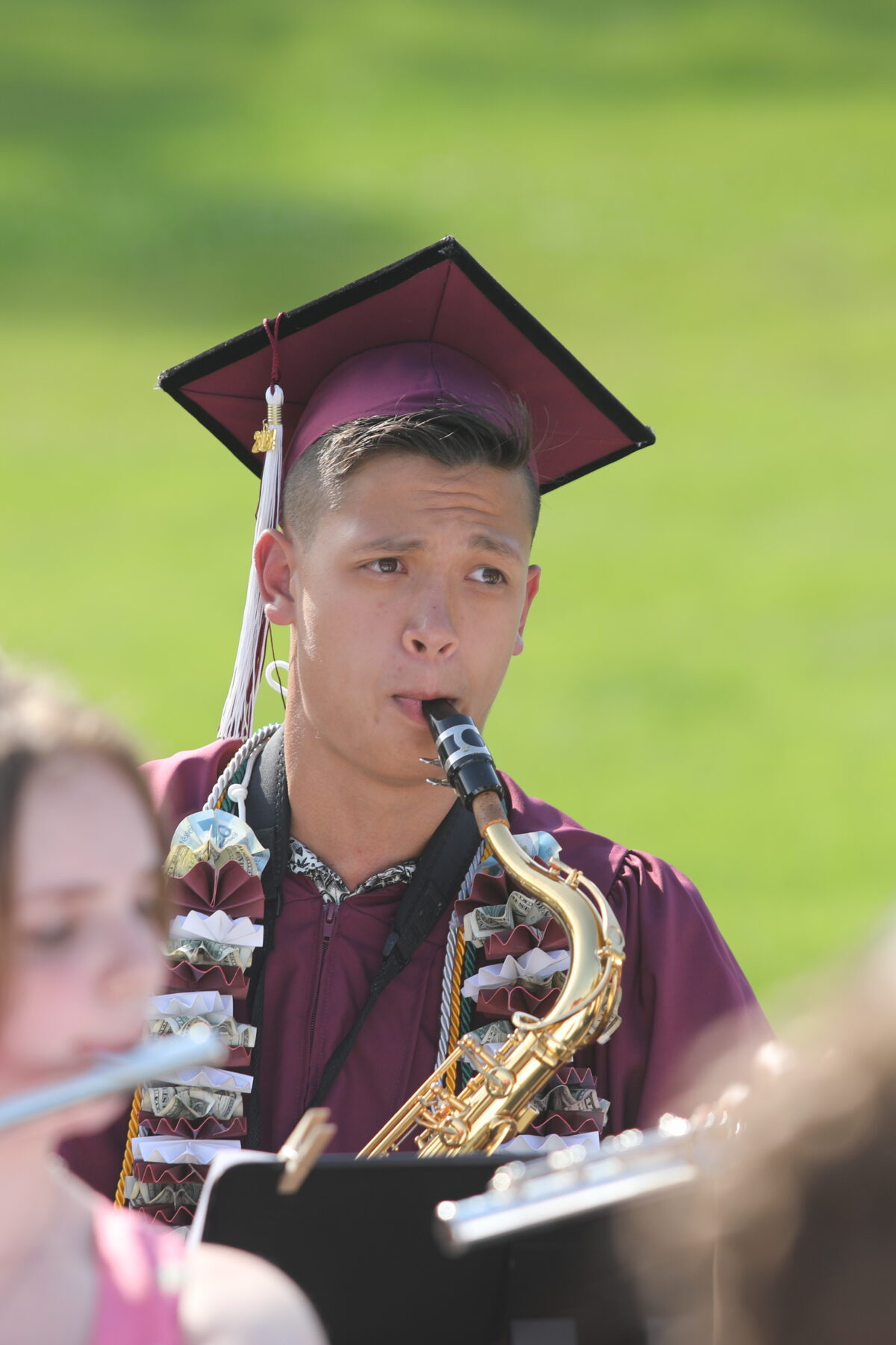 A person in a maroon graduation cap and gown plays a saxophone outdoors, accompanied by other musicians.