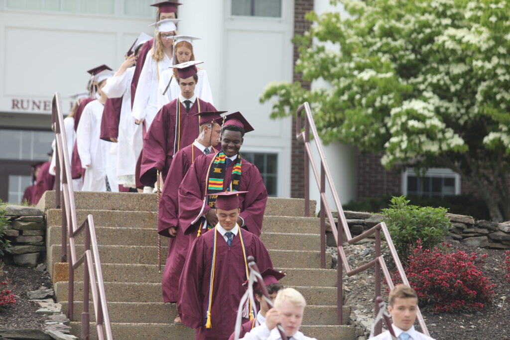 A line of students in graduation robes and caps walk down steps outside, with green bushes and a building in the background.
