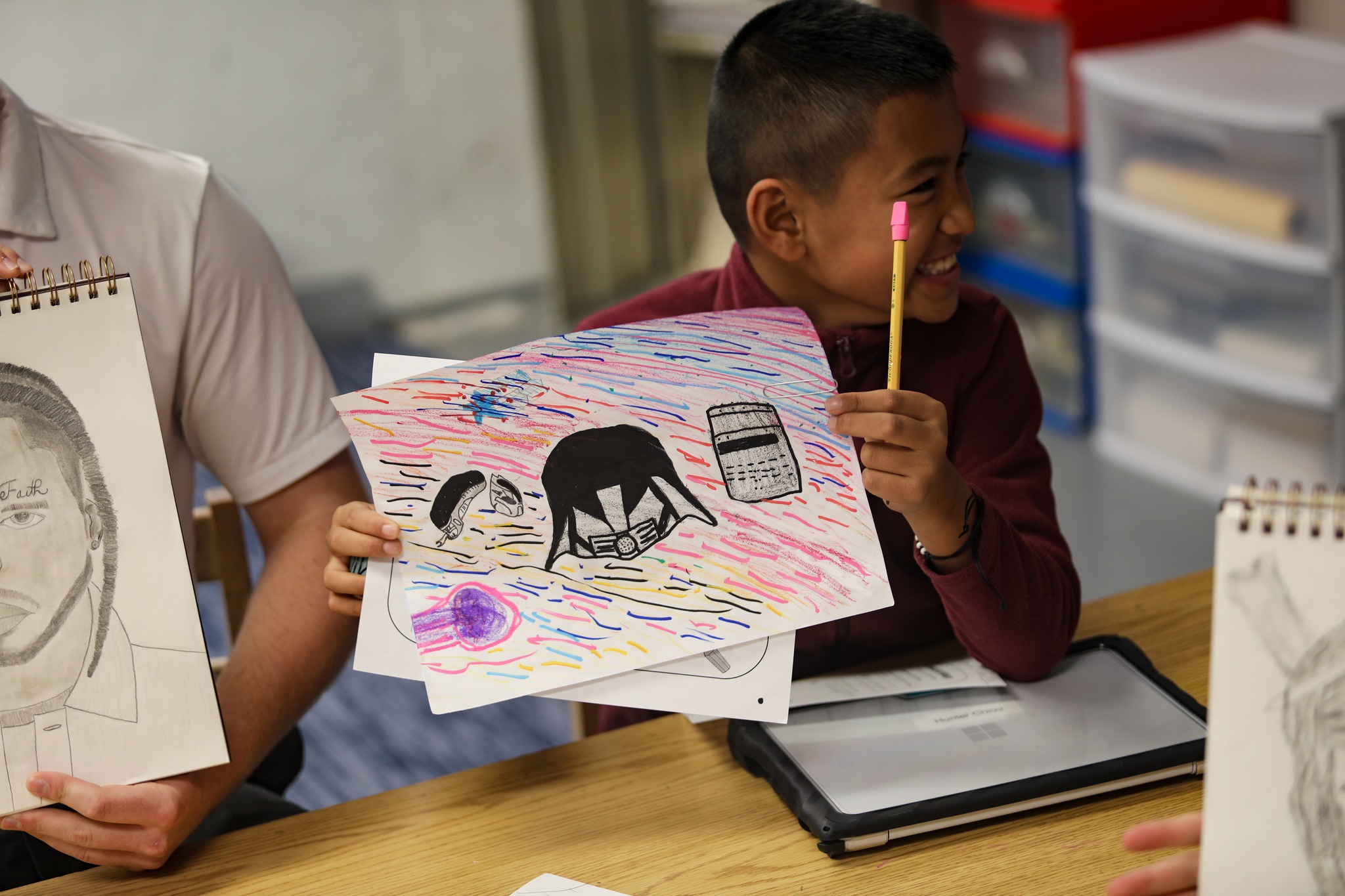 A smiling child holds up a colorful drawing in a classroom, sitting with others who also have sketchpads.