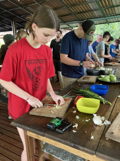 A group of people are chopping vegetables on wooden cutting boards in an outdoor kitchen. A girl in a red shirt and a boy in a blue shirt are at the forefront, focusing on their task.