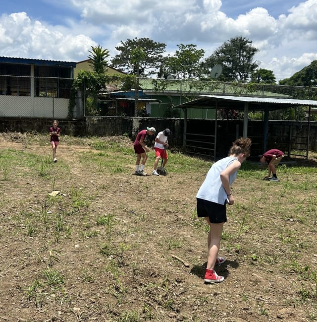 Children are outdoors in a grassy area near buildings, engaged in an activity that appears to require bending down and looking at the ground.