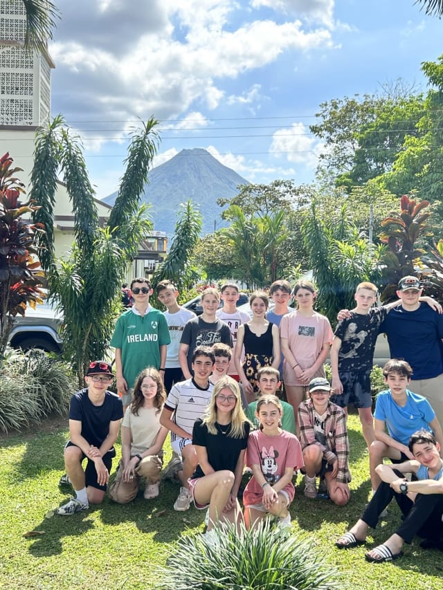 A group of young people stands and kneels on a lawn with a scenic view of a large mountain in the background. Trees and a building are also visible. They appear to be posing for the photograph.