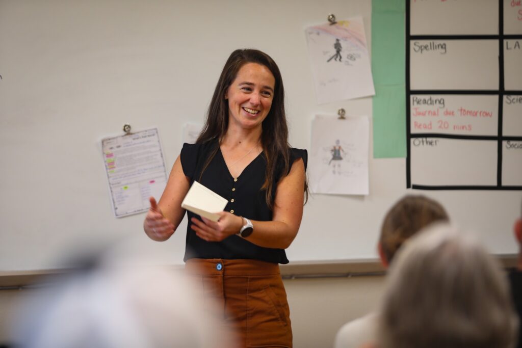 A woman stands and gestures while holding a notebook, smiling in front of a classroom whiteboard with various notes and papers attached.