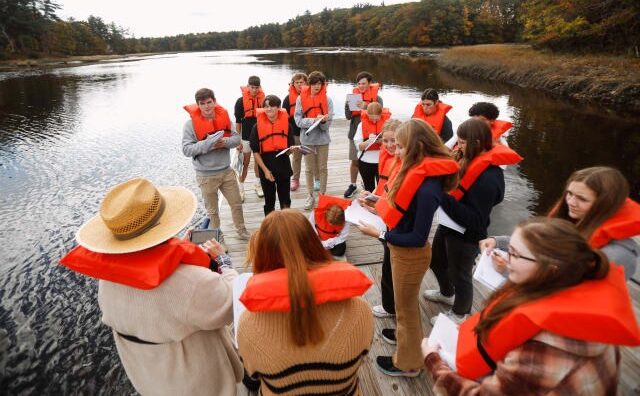 A group of people wearing orange life jackets stand on a dock by a lake, surrounded by autumn trees.