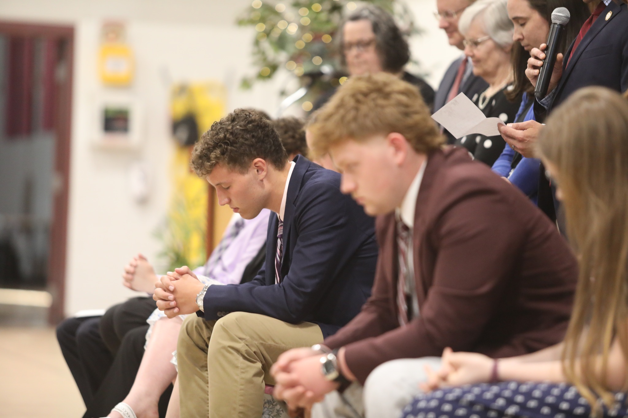 A group of four women stands outdoors, engaging with a person seated at a table. They are in a sunny area with trees and buildings in the background.