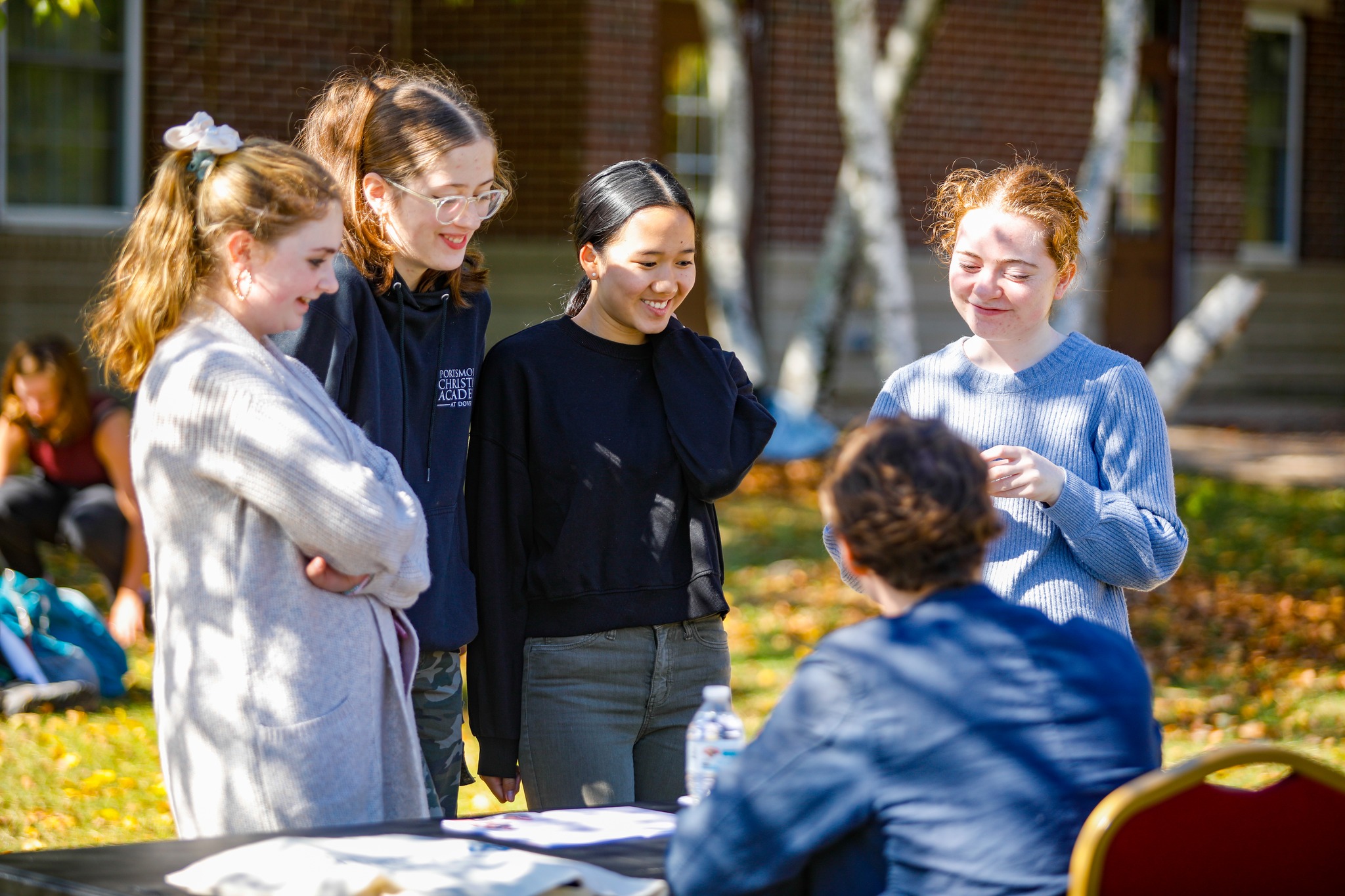 A group of four women stands outdoors, engaging with a person seated at a table. They are in a sunny area with trees and buildings in the background.