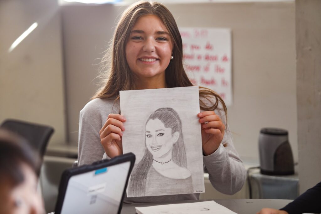A person holding a pencil drawing of someone with long hair at a desk, with a laptop and poster in the background.
