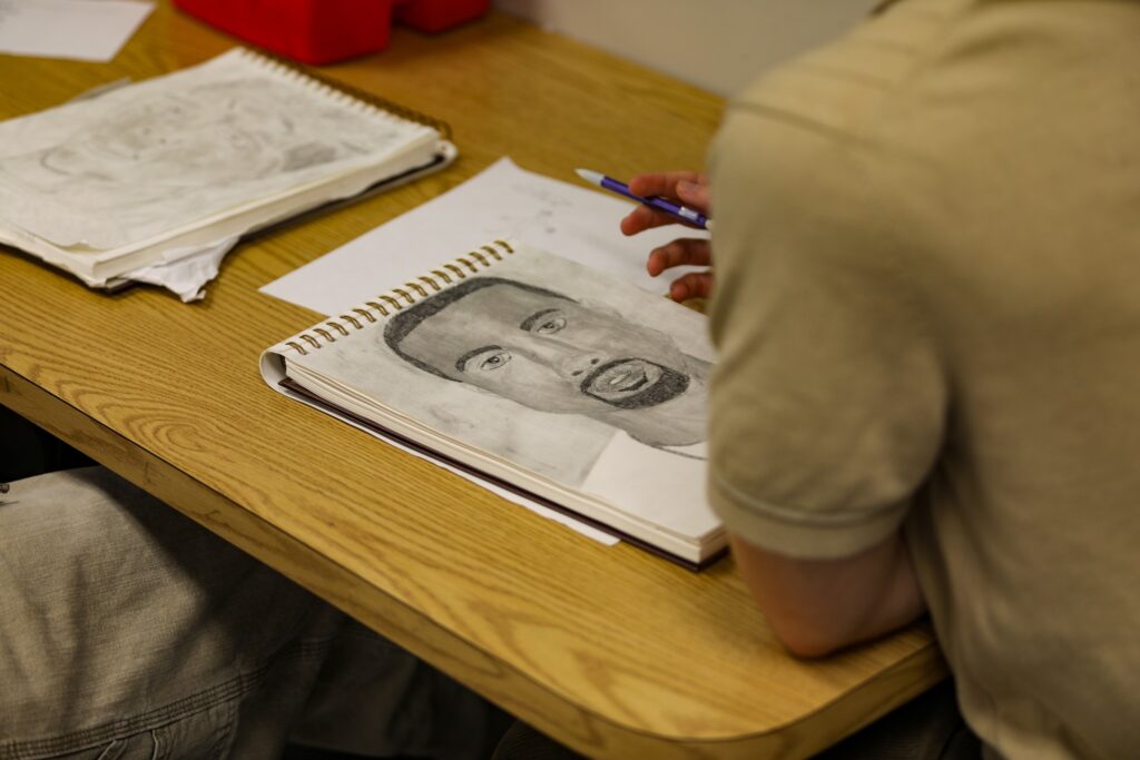 Person sketching a portrait on a drawing pad at a wooden table with additional sketches nearby.