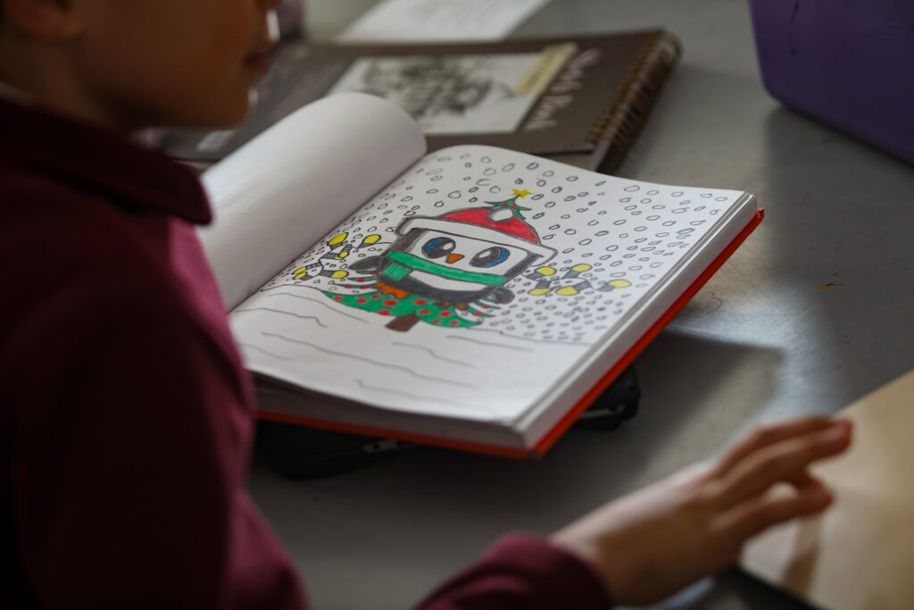 A child sits at a table with an open book featuring a colorful drawing of a festive character wearing a hat and scarf in a snowy scene.
