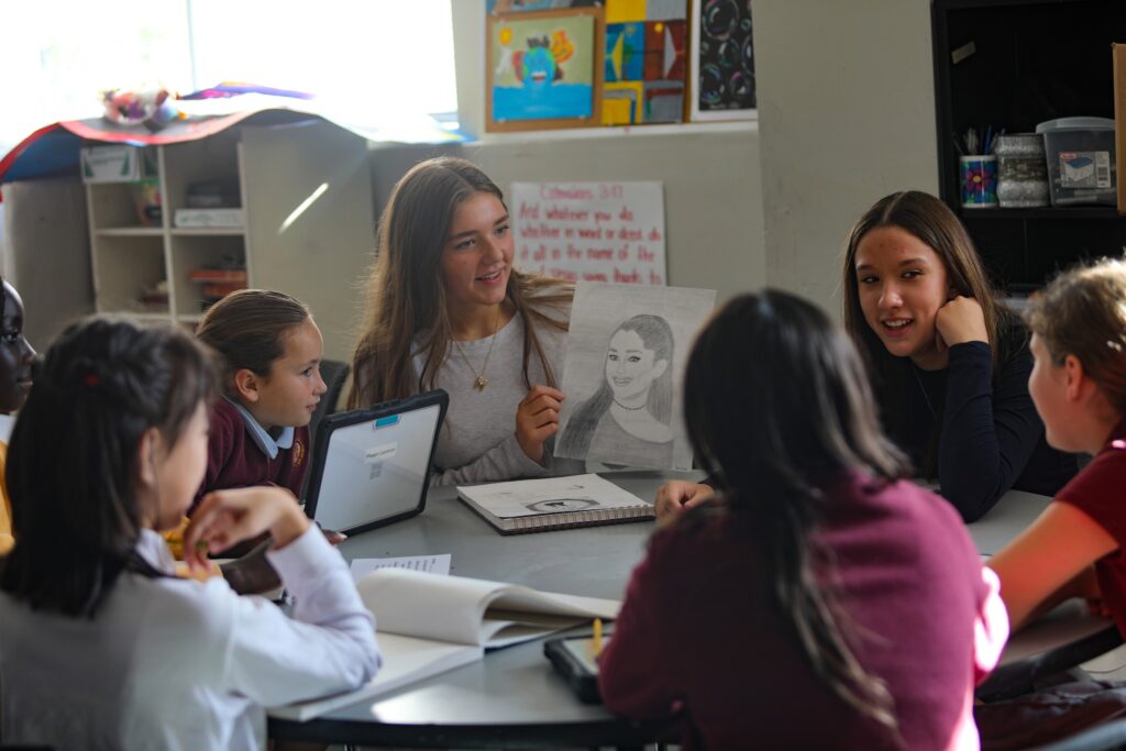 Students sit around a table in a classroom. One student holds up a drawing, while others look on attentively. Laptops and notebooks are on the table.