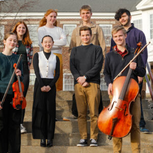 A group of eight young people stands on outdoor steps, some holding musical instruments like a violin and cello. They are in front of a brick building with large windows.