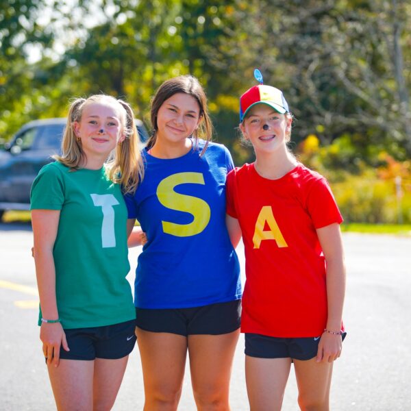 Three students in maroon graduation gowns and leis stand together indoors, smiling at the camera.