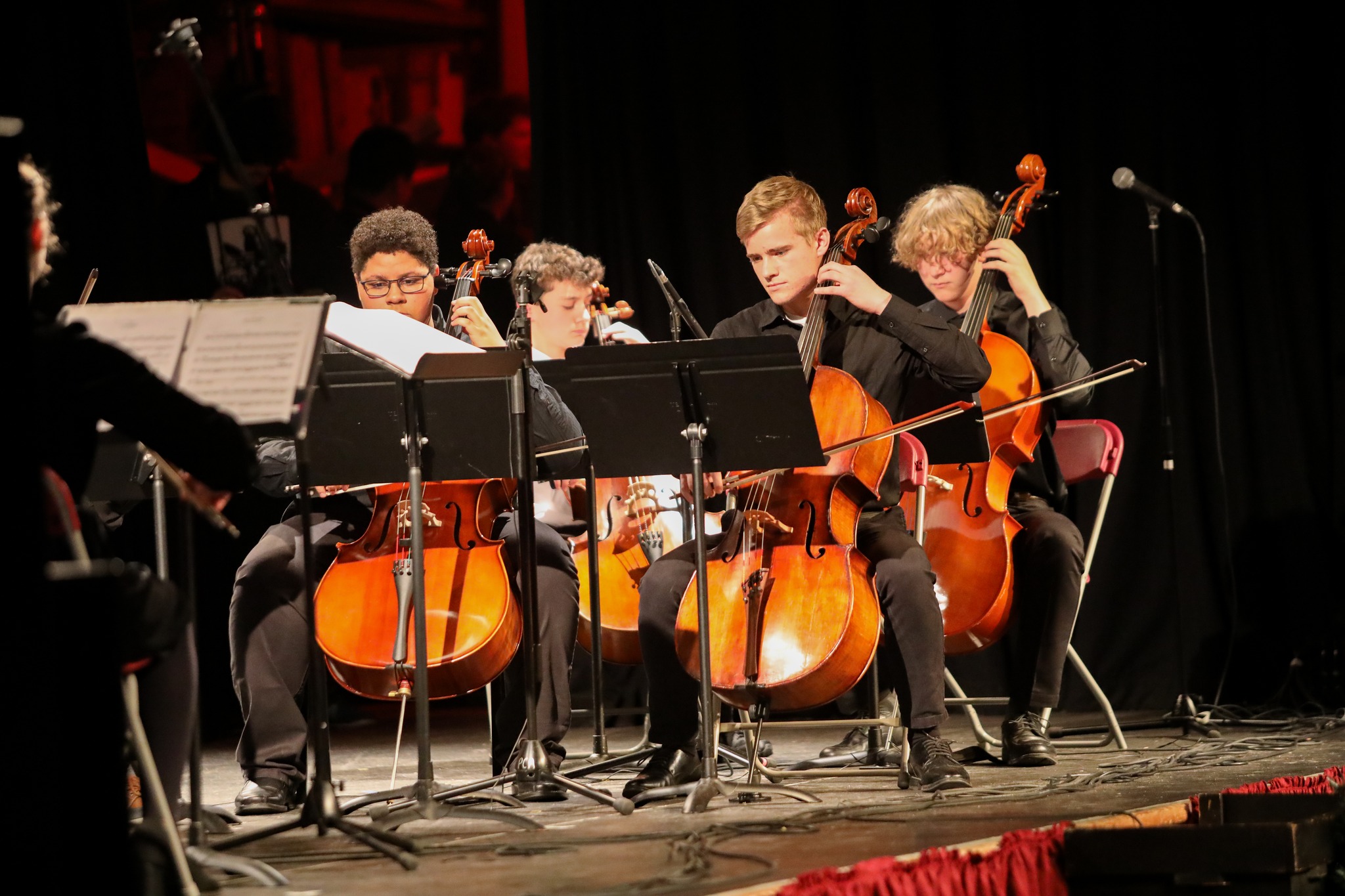 A group of musicians plays cellos on stage, seated in a row with music stands in front of them.