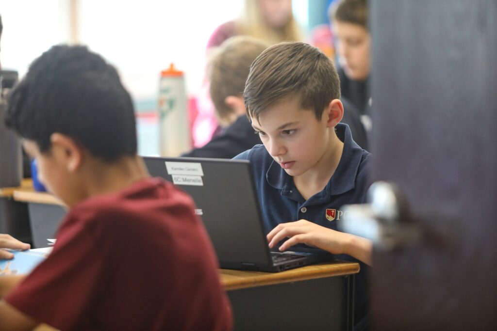 A student focused on typing on a laptop in a classroom, with other students working in the background.