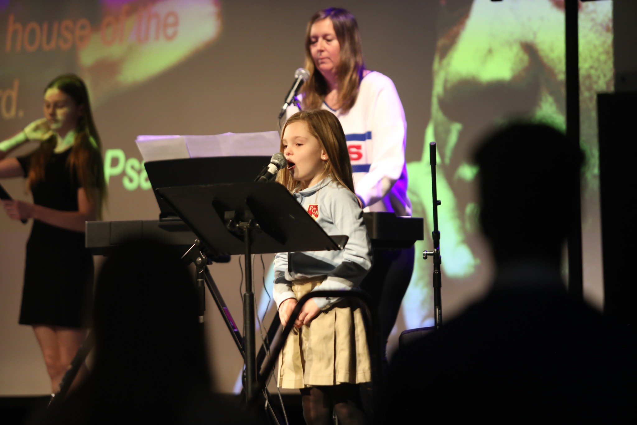 Young girl singing into a microphone on stage, accompanied by a woman playing a keyboard. Another woman stands beside them, with an audience silhouette in the foreground.