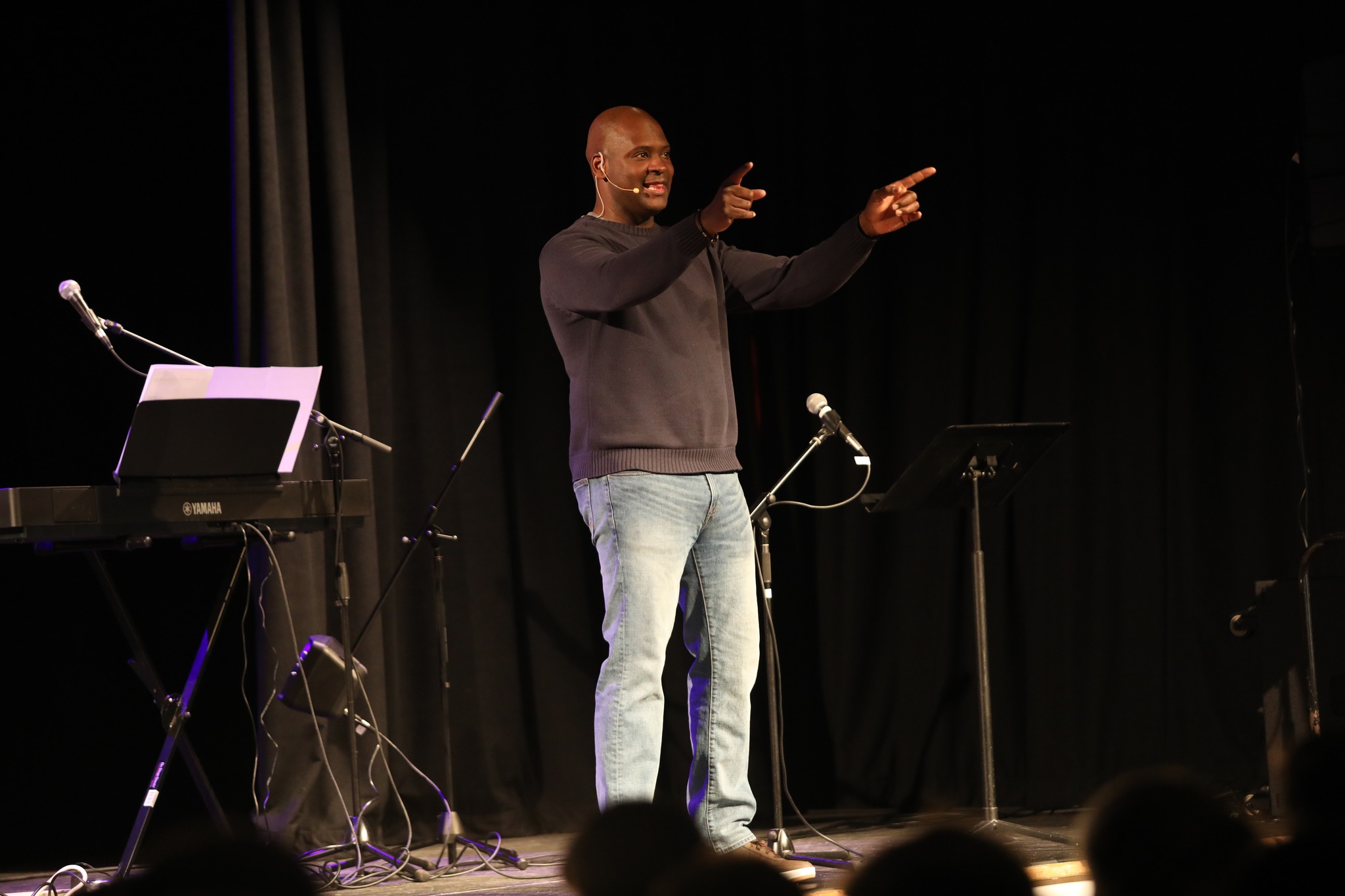 A person stands on stage pointing and smiling, with microphones and a keyboard nearby. The background is dark.