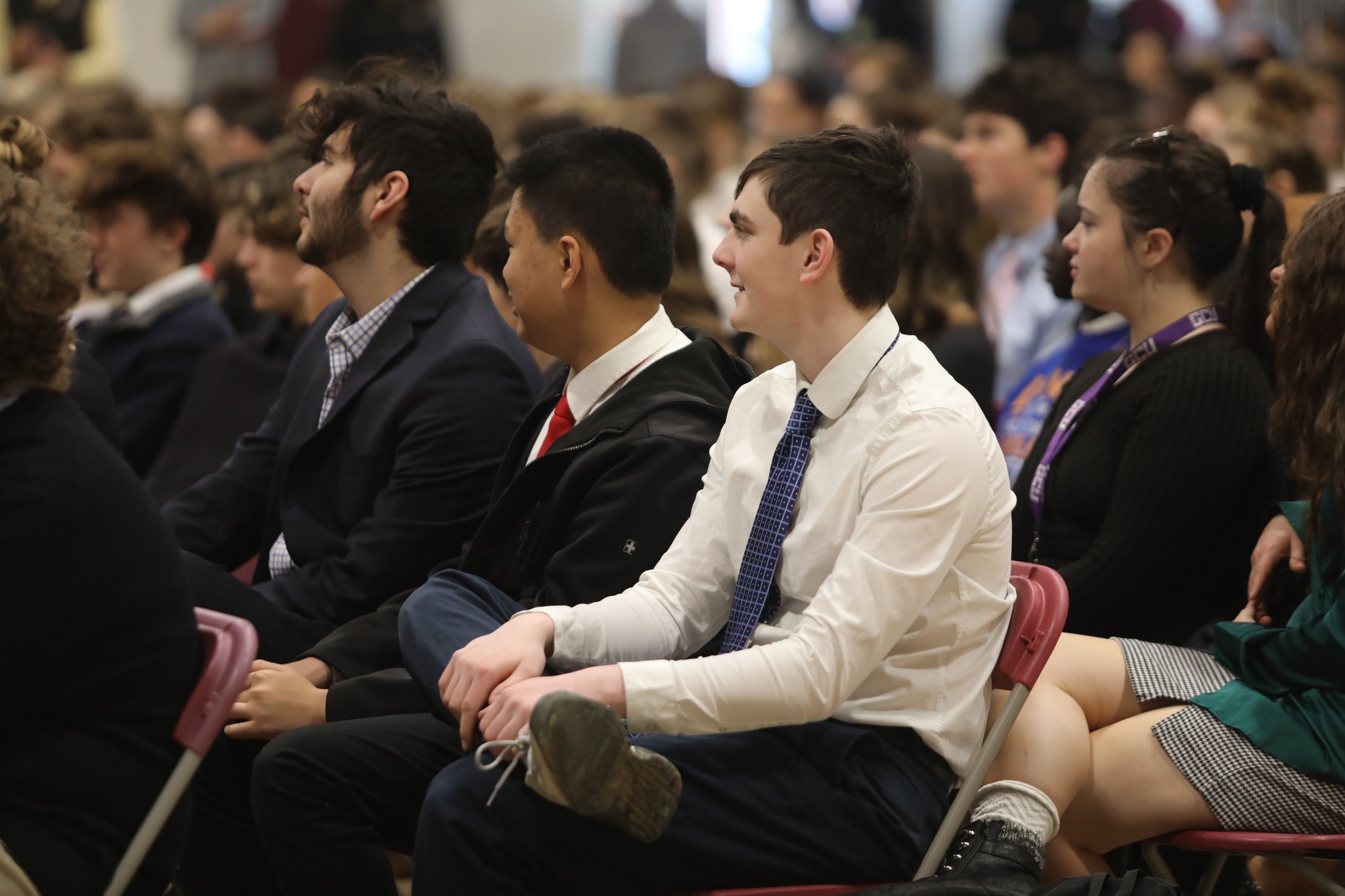 A group of people seated in rows at an indoor event, listening attentively. Some are dressed in formal attire.