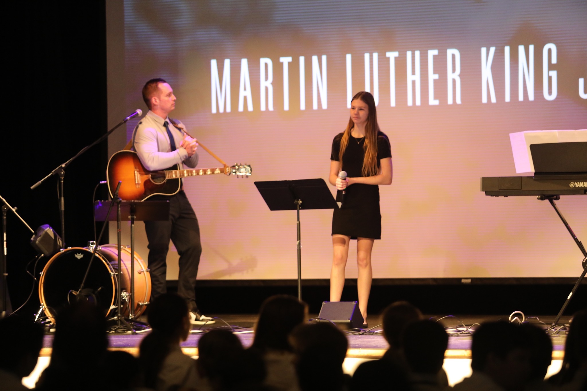 Two musicians on stage, one holding a guitar and the other a microphone, with a backdrop displaying "Martin Luther King Jr.