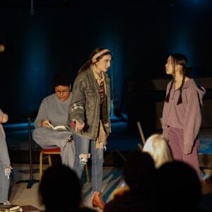 Four people on stage in casual attire, with one sitting and reading while two stand and converse. The background is dark, and a small audience is visible in the foreground.