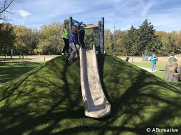 Three children stand at the top of a playground slide set on a grassy hill, with trees and people visible in the background.