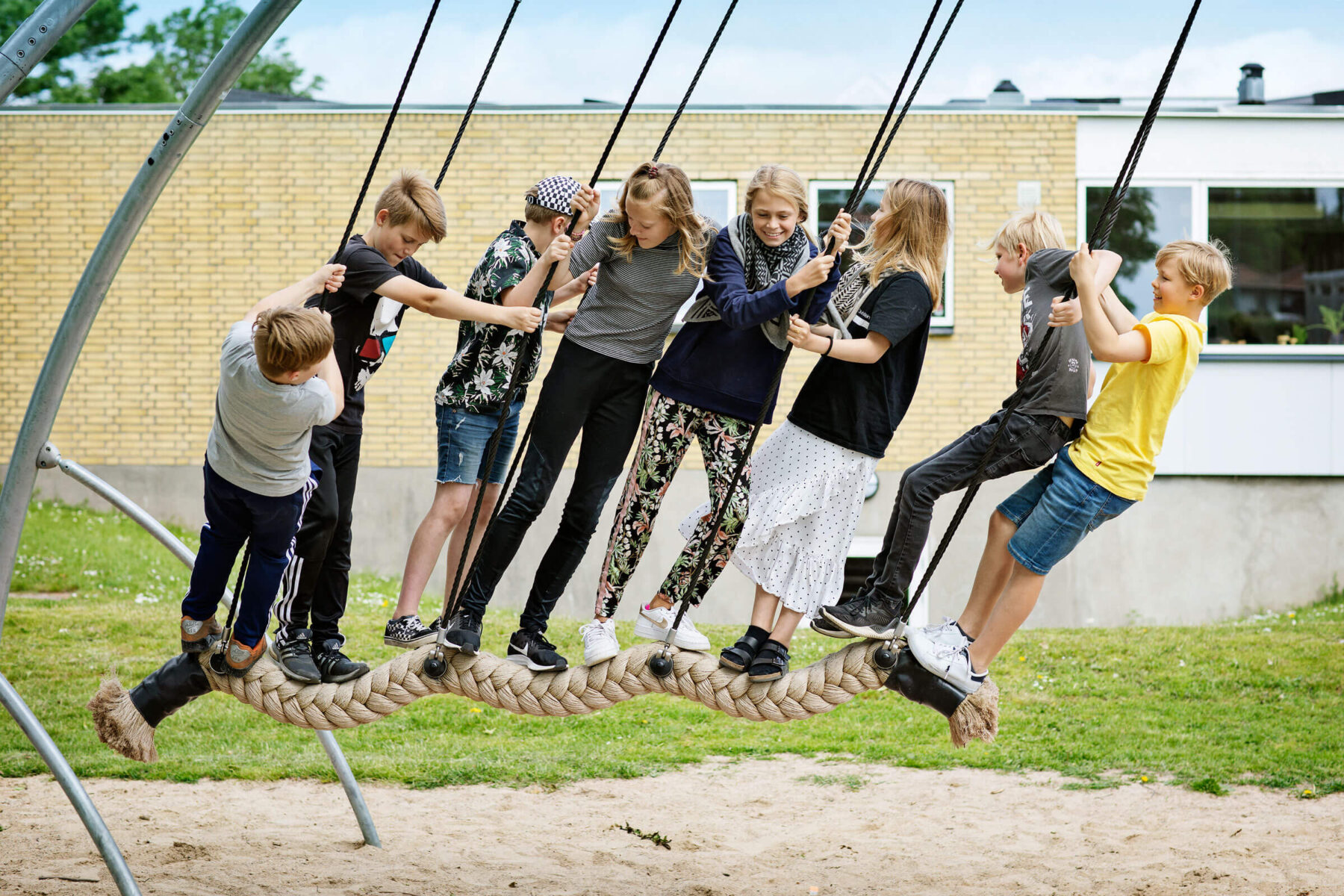 Seven children stand together on a rope swing at a playground, balancing as they hold onto ropes, with a building and grass in the background.