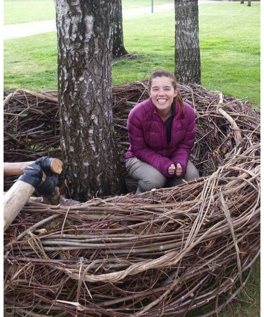 A woman in a purple jacket smiles while sitting inside a large nest made of intertwined branches at the base of two trees in a grassy park.