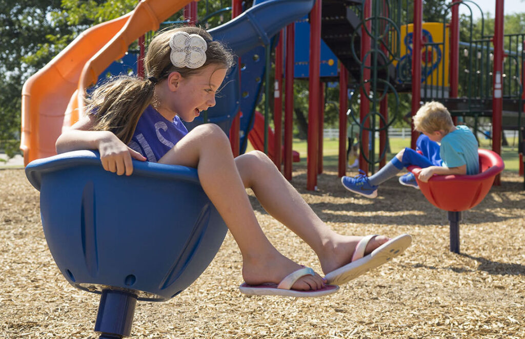 Two children play separately on spinning playground seats, with slides and climbing equipment in the background on a sunny day.