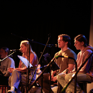A group of young people sit on stools on a stage, each with a microphone and sheet music, appearing to sing or read aloud.