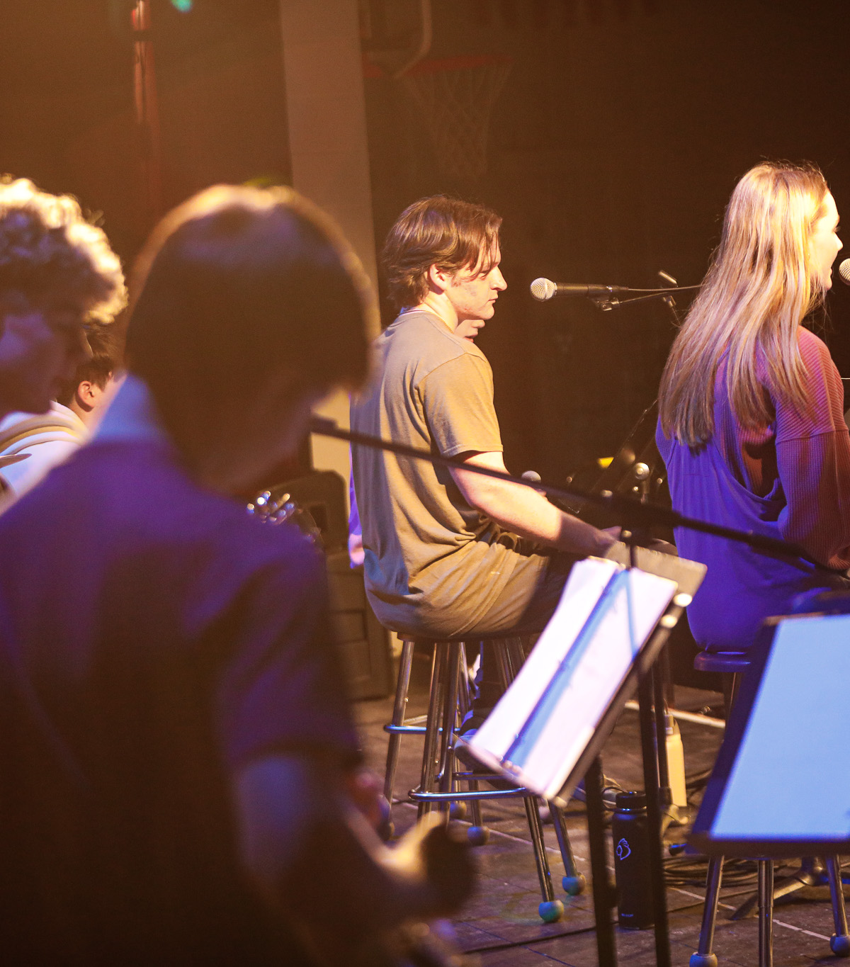 Several musicians sit on stools facing microphones and music stands on a dimly lit stage, performing or rehearsing together.