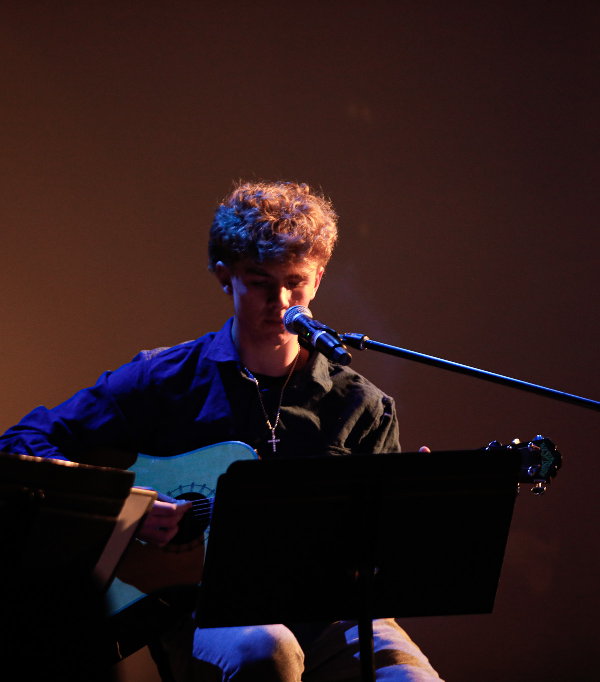 A person sits on stage playing an acoustic guitar and singing into a microphone under warm stage lighting, with a music stand in front of them.