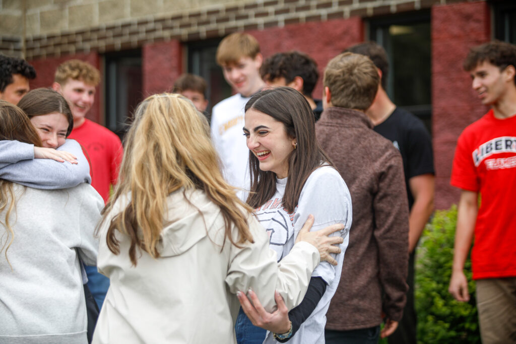 A group of young people stands outside a brick building. One woman in the foreground smiles as she is greeted by another woman. Several people interact in the background.