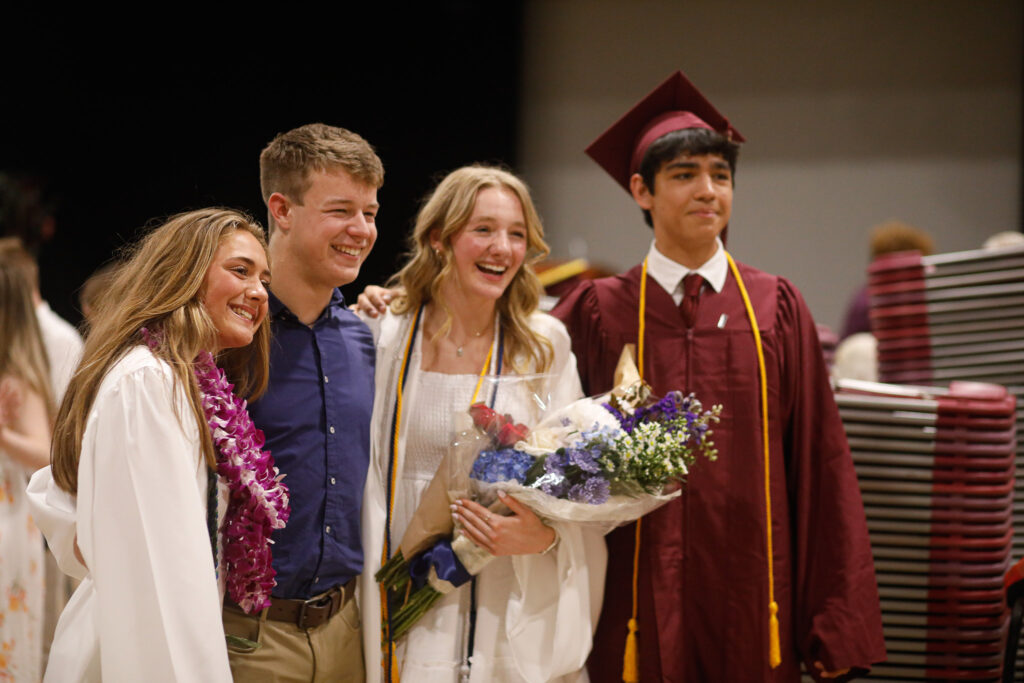 Four graduates pose together, two in white gowns and one in a maroon cap and gown, smiling and holding flowers at a graduation ceremony.
