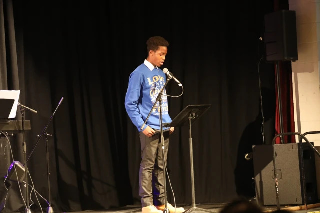 A young man stands on stage reading from a music stand into a microphone, with a black curtain backdrop and stage equipment nearby.
