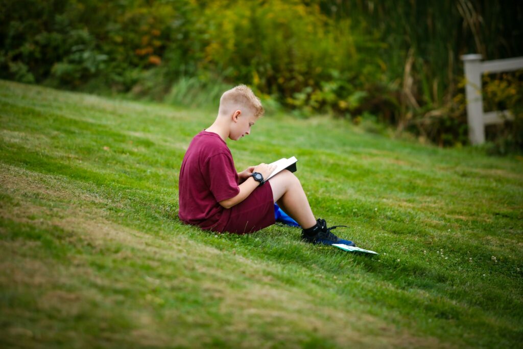 A boy sits on a grassy slope outdoors, wearing a maroon shirt and shorts, reading a book with an umbrella beside him. Shrubs and a wooden fence are in the background.