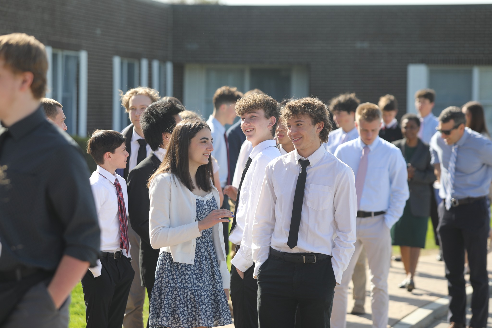 A group of young people dressed in semi-formal attire stand and converse outside a brick building on a sunny day.
