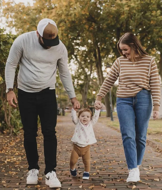 A man and woman hold hands with a smiling toddler walking between them on a leaf-covered path in a park, with trees in the background.
