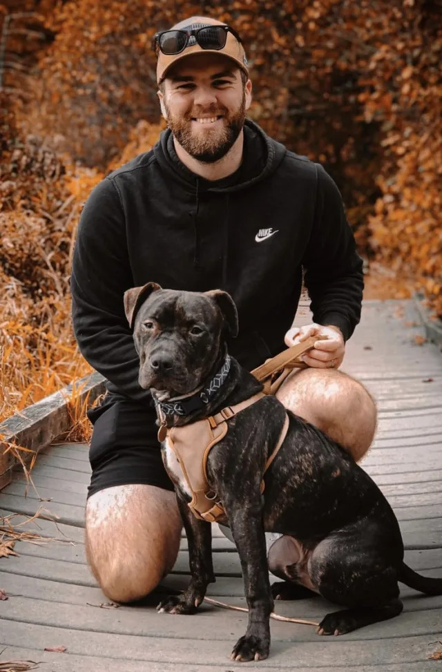 A man in a black hoodie and shorts kneels on a wooden path outdoors, smiling at the camera while holding a black and brown dog on a leash. Autumn foliage surrounds them.