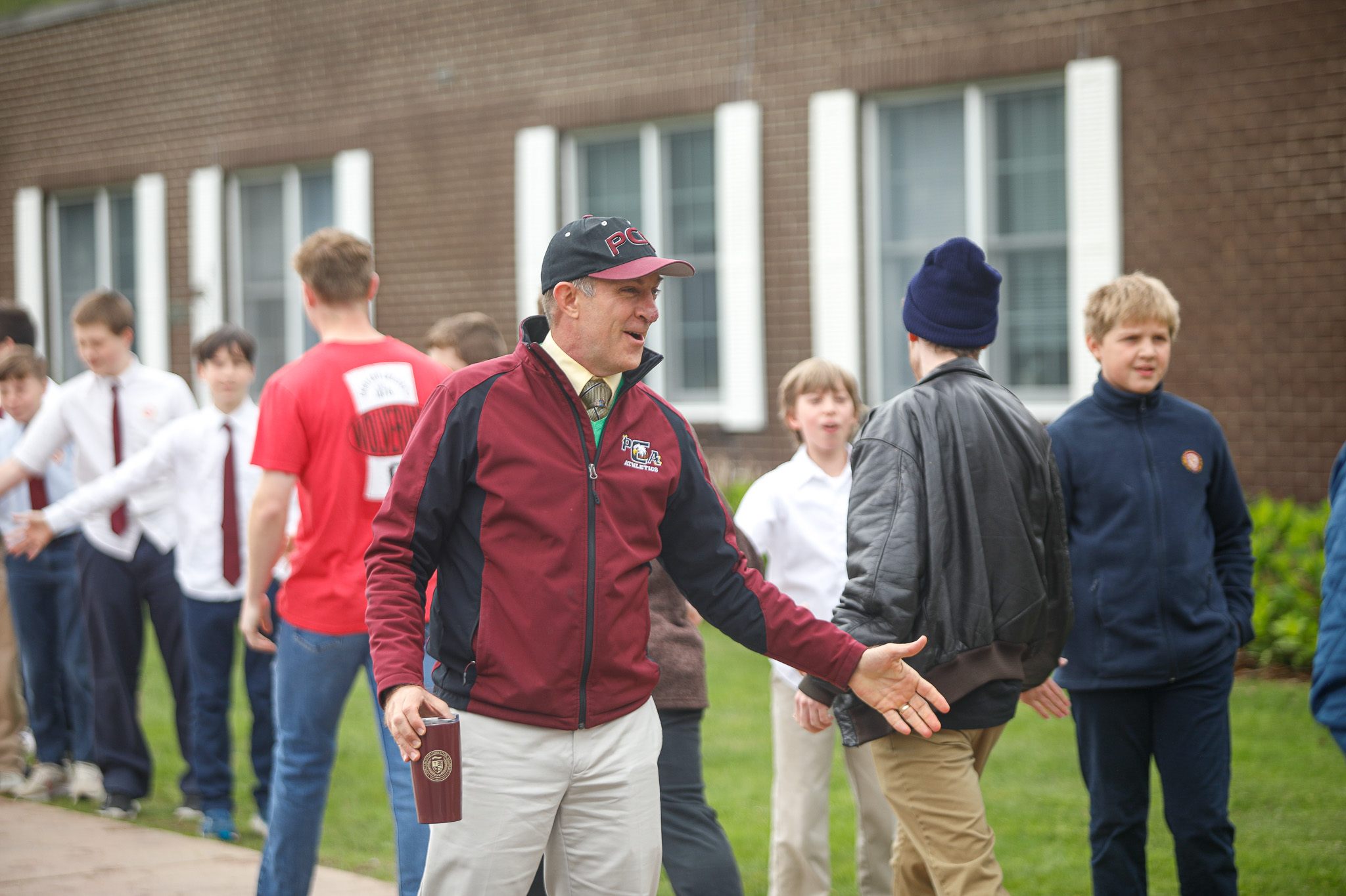 A man in a maroon jacket and cap high-fives students standing in a line outside a brick building.