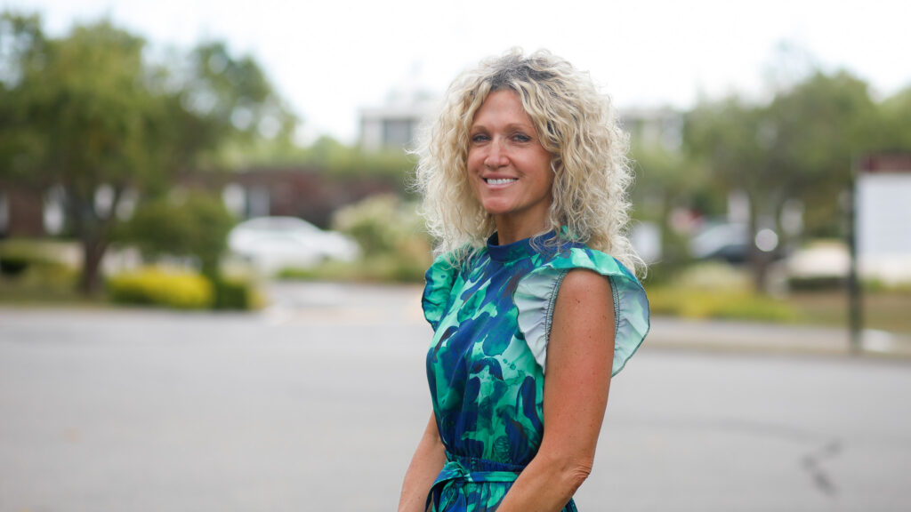 A woman with curly blonde hair wearing a blue and green patterned dress stands outdoors in a parking lot, smiling at the camera. Trees and blurred buildings are in the background.