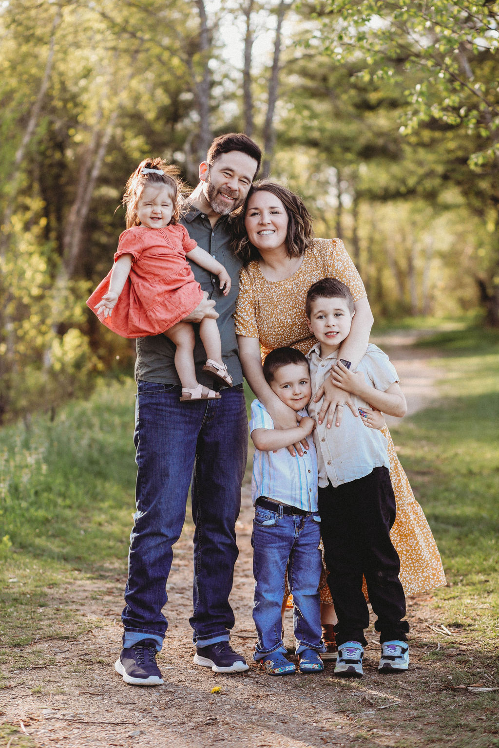 A family of five poses on a wooded path; two adults stand close together with a girl in an orange dress, while two young boys stand in front, all smiling.