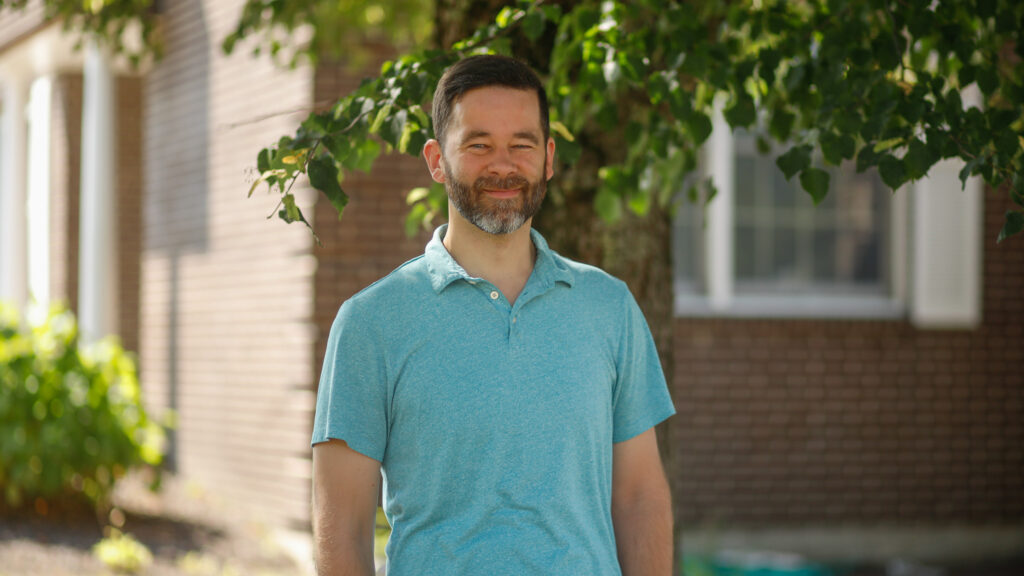 A man with short dark hair and a beard stands outside in front of a tree, wearing a light blue polo shirt, with a brick building in the background.